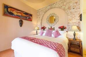 A bedroom with a brick wall, a large bed with pink bedding, a painting, a mirror, and two bedside lamps. - Casa Kukana- 4 bedrooms - sleeps 9 (Sayulita)