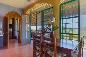 A dining area with a wooden table and chairs, large windows with green frames, and a view of the outdoors. - Casa Kukana- 4 bedrooms - sleeps 9 (Sayulita)
