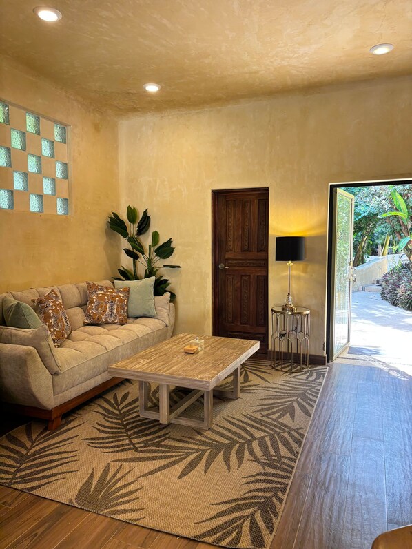 A living room with a sofa, coffee table, and a door leading to a garden. - Romantic Jungle Bungalow w/ Pool Near Town & Beach (Sayulita)