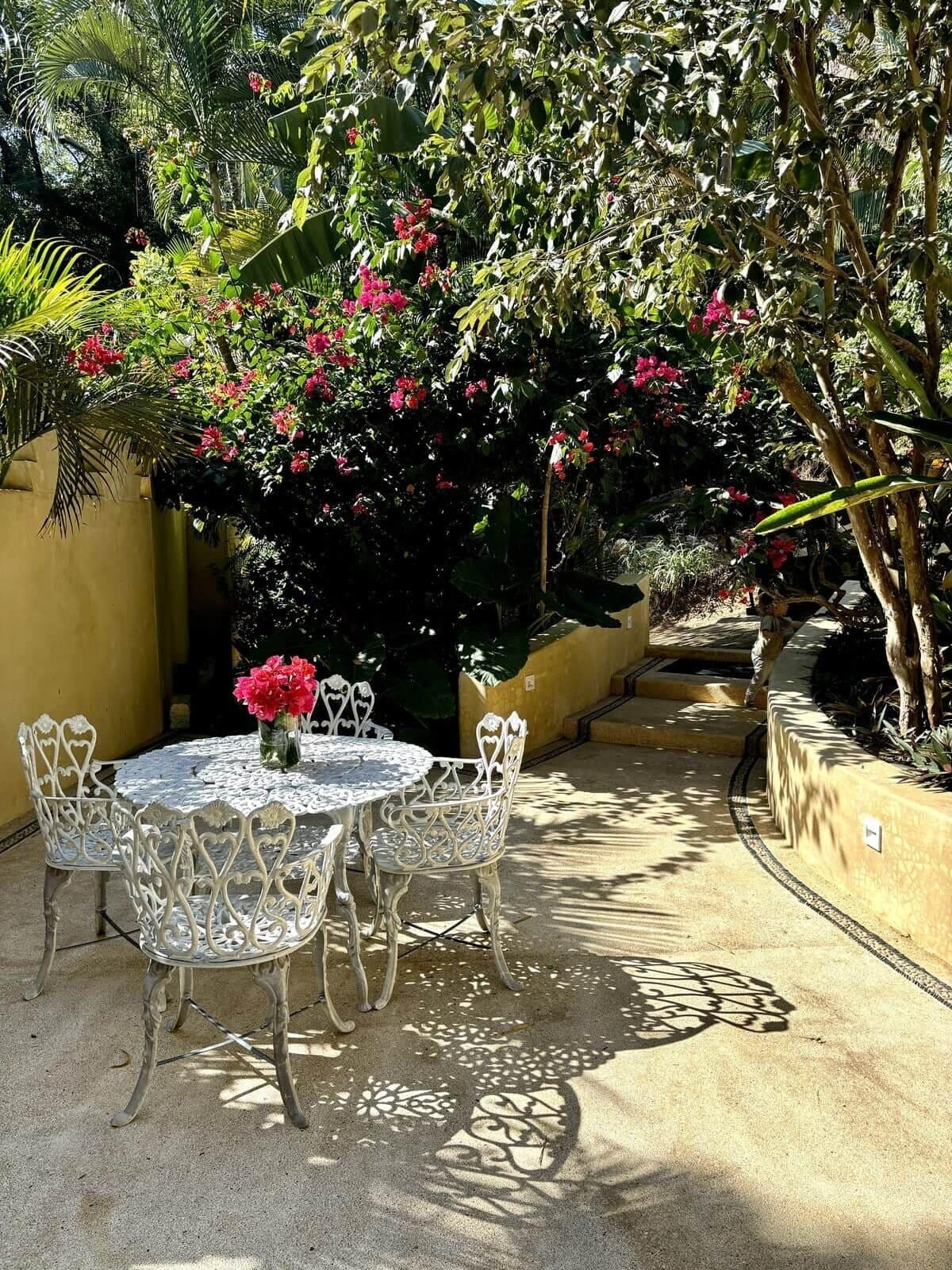 Une terrasse extérieure avec une table ronde et des chaises, entourée d’une végétation luxuriante et de plantes en fleurs.