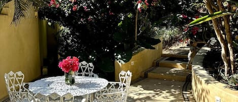 Une terrasse extérieure avec une table ronde et des chaises, entourée d’une végétation luxuriante et de plantes en fleurs.
