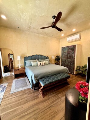 A bedroom with a bed, a ceiling fan, a nightstand, a lamp, a mirror, a wall-mounted air conditioner, and a decorative panel. - Romantic Jungle Bungalow w/ Pool Near Town & Beach (Sayulita)