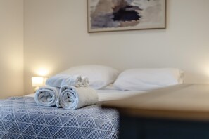 Ein ordentlich bezogenes Bett mit weißen Leinen, einer gemusterten Tagesdecke und einem aufgerollten Handtuch daneben.