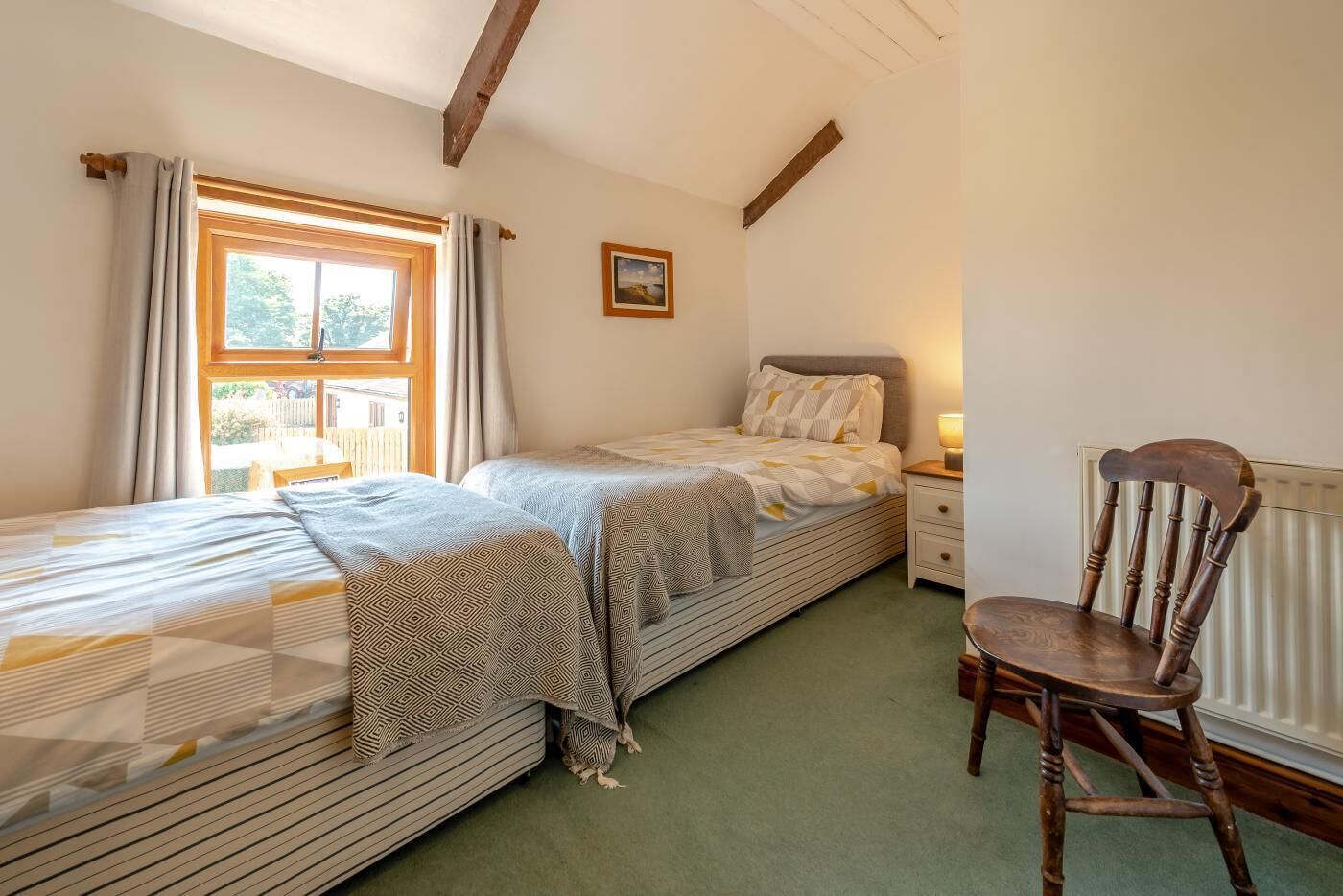 Une chambre avec deux lits, une chaise, une table de chevet avec une lampe, une fenêtre avec des rideaux et un tableau au mur.