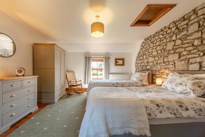 Een slaapkamer met een stenen muur, een bed met bloemenbedekking, een dressoir met laden, een stoel en een raam met gordijnen.