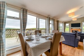A dining area with a table set for four, a sofa, and a television in a room with large windows. - 2 Bed in Glenluce (oc-c36927) (Glenluce)