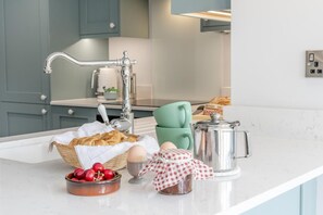 A kitchen counter with a silver faucet, a basket of bread, a bowl of cherries, a coffee pot, and a green chair. - 2 Bed in Tenby (oc-b41400) (Tenby)