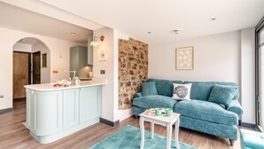 A modern living room with a teal sofa, a small white table, and a kitchen area with a light blue cabinet. - 2 Bed in Tenby (oc-b41400) (Tenby)