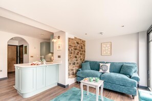 A modern living room with a teal sofa, a small white table, and a kitchen area with a light blue cabinet. - 2 Bed in Tenby (oc-b41400) (Tenby)