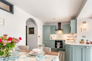 A modern kitchen with teal cabinetry, a central island, and a dining area with a table set for two. - 2 Bed in Tenby (oc-b41400) (Tenby)