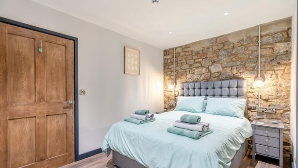 A bedroom with a stone wall, a bed with pillows, a bedside table, and a wooden door. - 2 Bed in Tenby (oc-b41400) (Tenby)