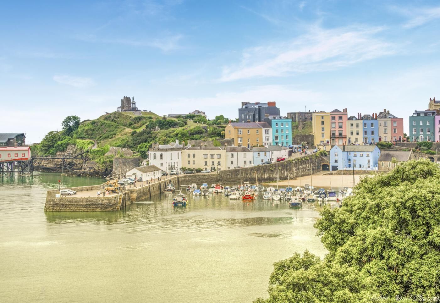 A coastal town with colorful buildings, a harbor with boats, and a hill with a structure on top.