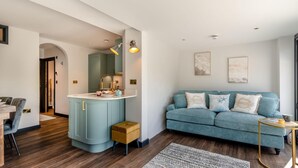 A modern living room with a blue sofa, a small wooden stool, and a dining area with a table and chairs. - 2 Bed in Tenby (oc-b41402) (Tenby)