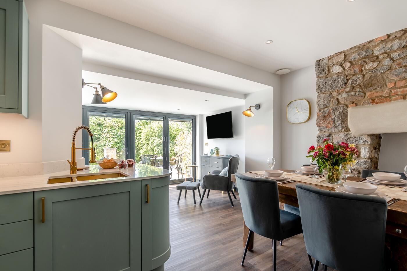 A modern kitchen with a curved island, a dining area with a stone accent wall, and a large window providing a view of greenery.