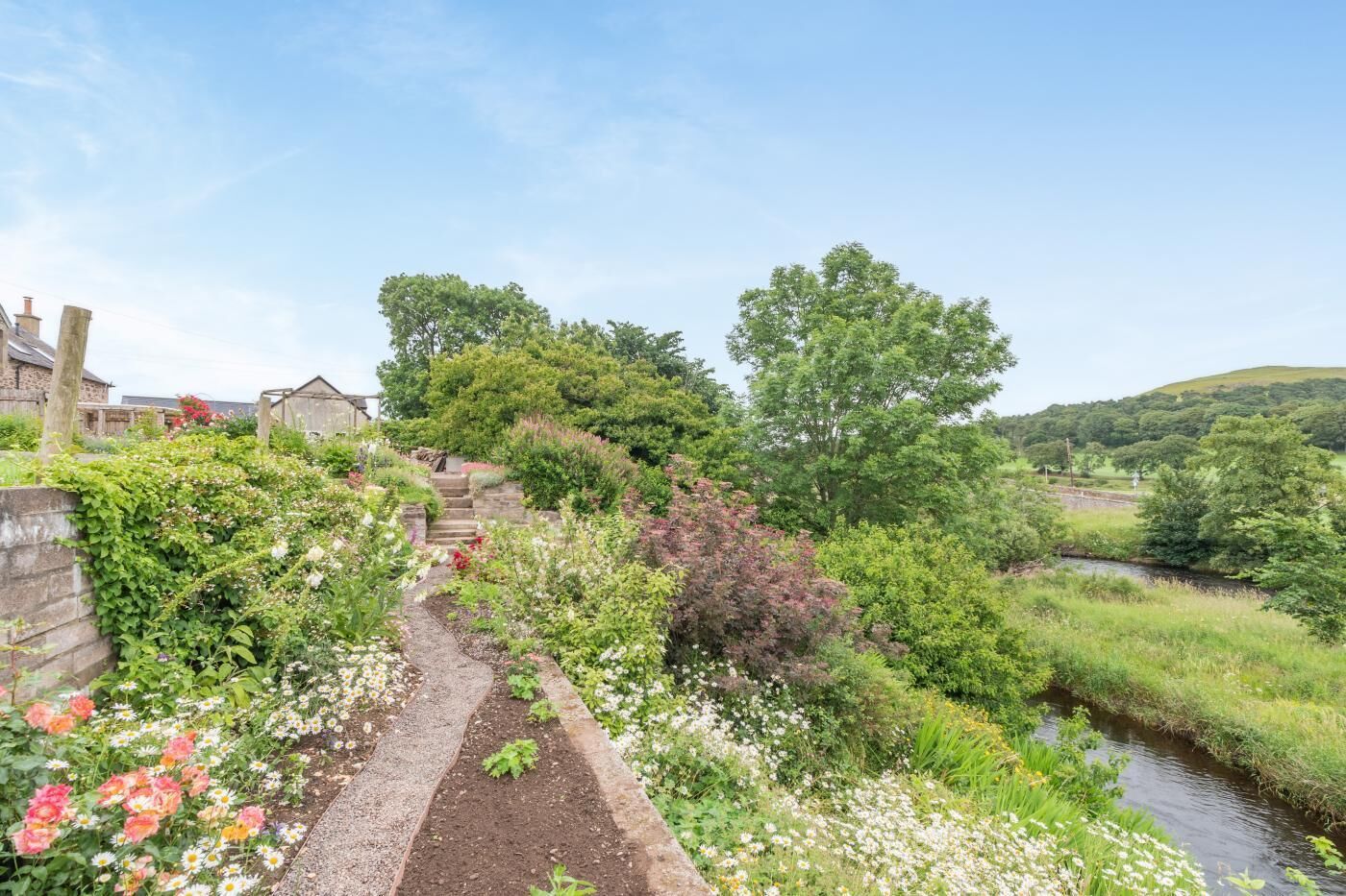 Ein Gartenweg führt zu einem Haus mit Blick auf einen Fluss und Hügel im Hintergrund.