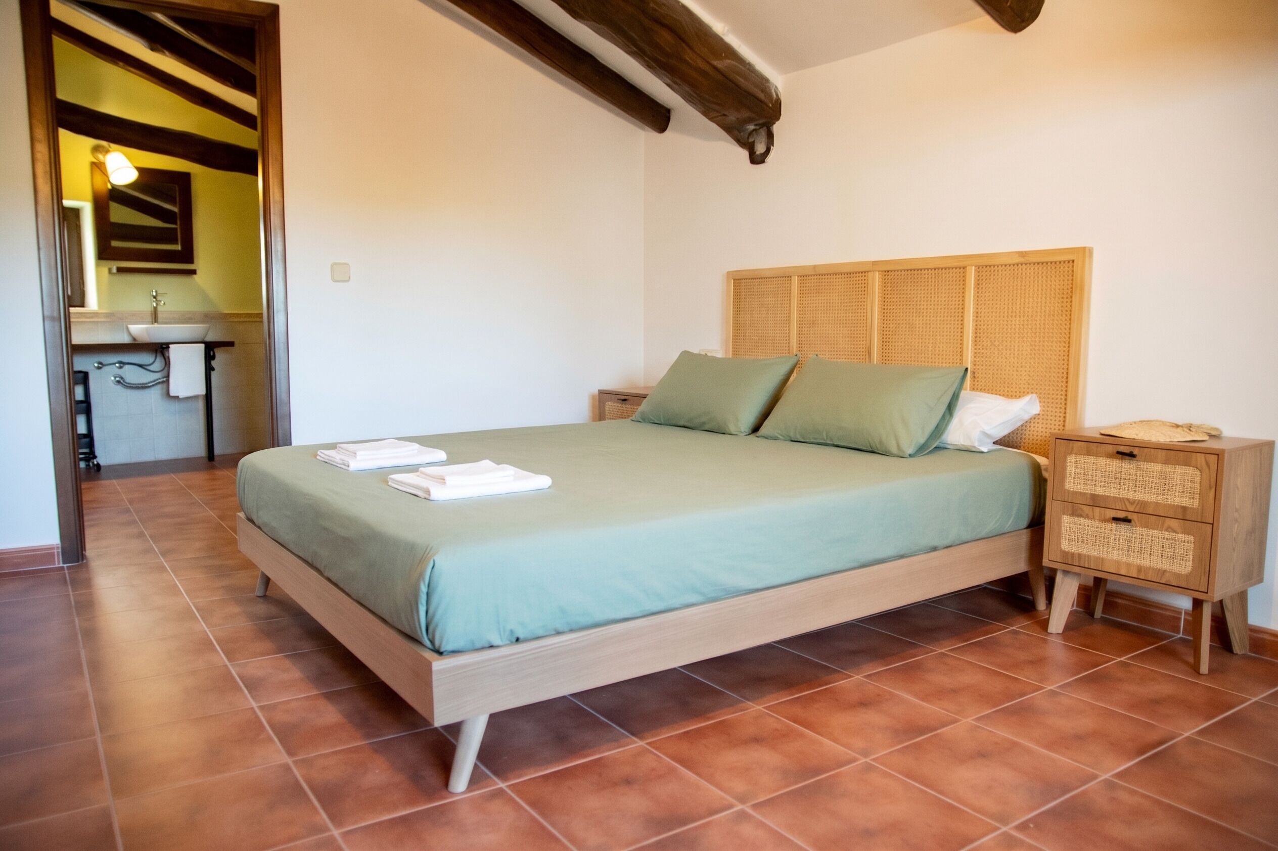 Un dormitorio con cama, mesitas de noche, un tablero auxiliar, cabecera de madera y un baño visible a través de una puerta abierta.
