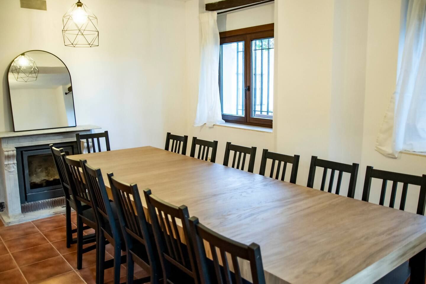 A dining room with a large wooden table and chairs, a fireplace, and a window with curtains.