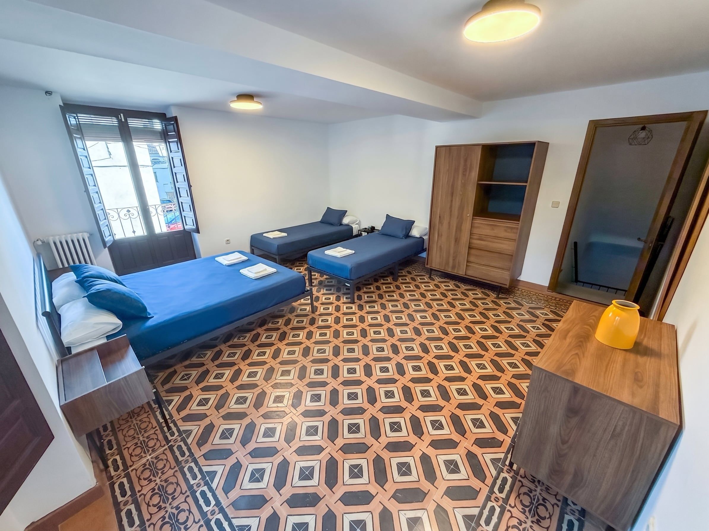 Habitación pequeña y funcional con dos camas individuales, un armario de madera, una mesita y un radiador.