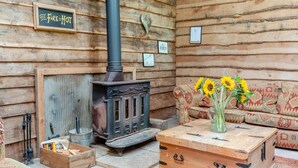 Interior rústico de una cabaña con estufa de leña, un sofá con cojines estampados y un jarrón con girasoles sobre una mesa de madera.