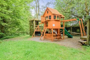 Ein hölzernes Spielhaus mit Rutsche und Kletterbereich, umgeben von Bäumen und Gras.