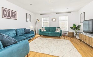 Ein Wohnzimmer mit zwei smaragdgrünen Sofas, einem weißen Teppich, einer Stehlampe und einem Fernseher auf einem Holzschrank.