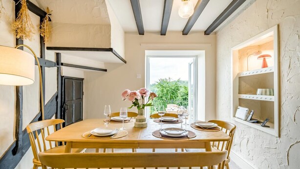 Een eetgedeelte met een houten tafel gedekt voor vier personen, een vaas met bloemen en uitzicht op groen buiten.