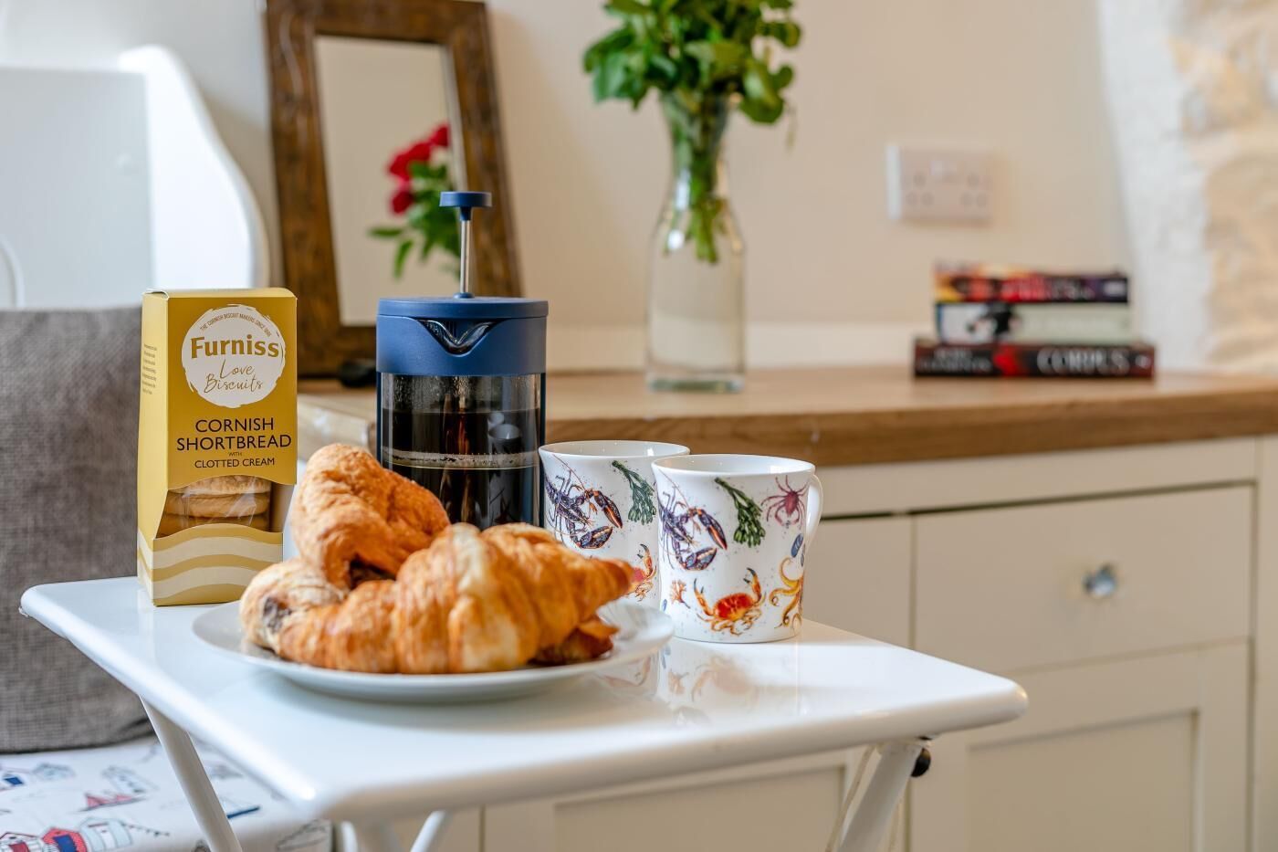 A breakfast setup with a box of Cornish Shortbread biscuits, croissants, a coffee maker, and two patterned mugs on a white table.