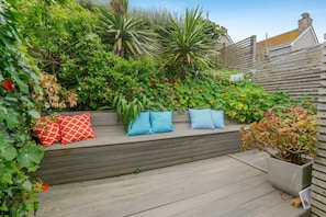 A wooden deck with a bench and cushions, surrounded by lush greenery and a variety of plants. - 3 Bed in St. Ives (oc-d42669) (St Ives)