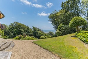 Un jardin aménagé avec un chemin de gravier menant à un bâtiment, entouré d’une végétation luxuriante et offrant une vue sur la mer.