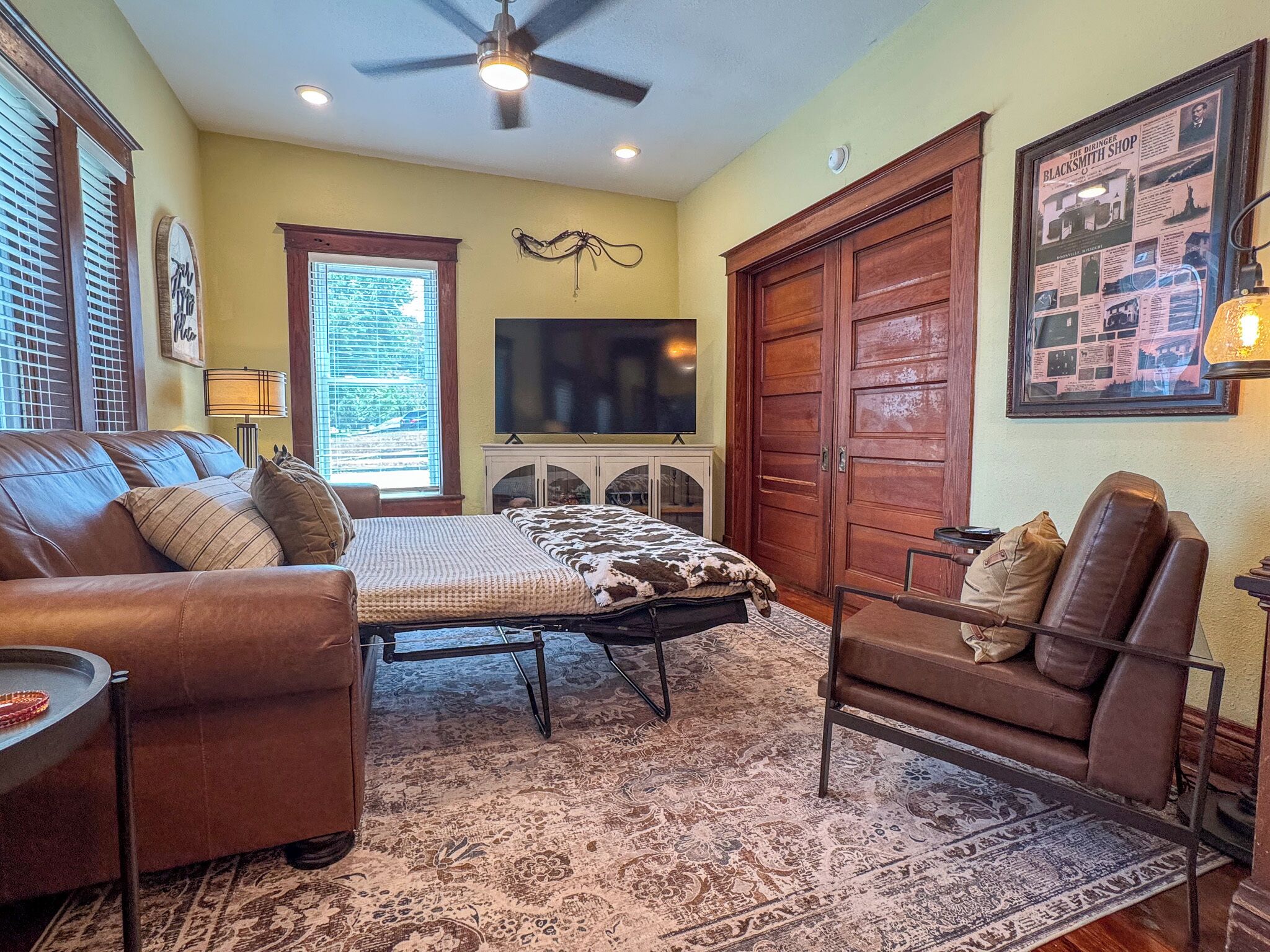 A room with a brown leather sofa, a bed with a patterned bedspread, a chair, a television, and a large wooden door.