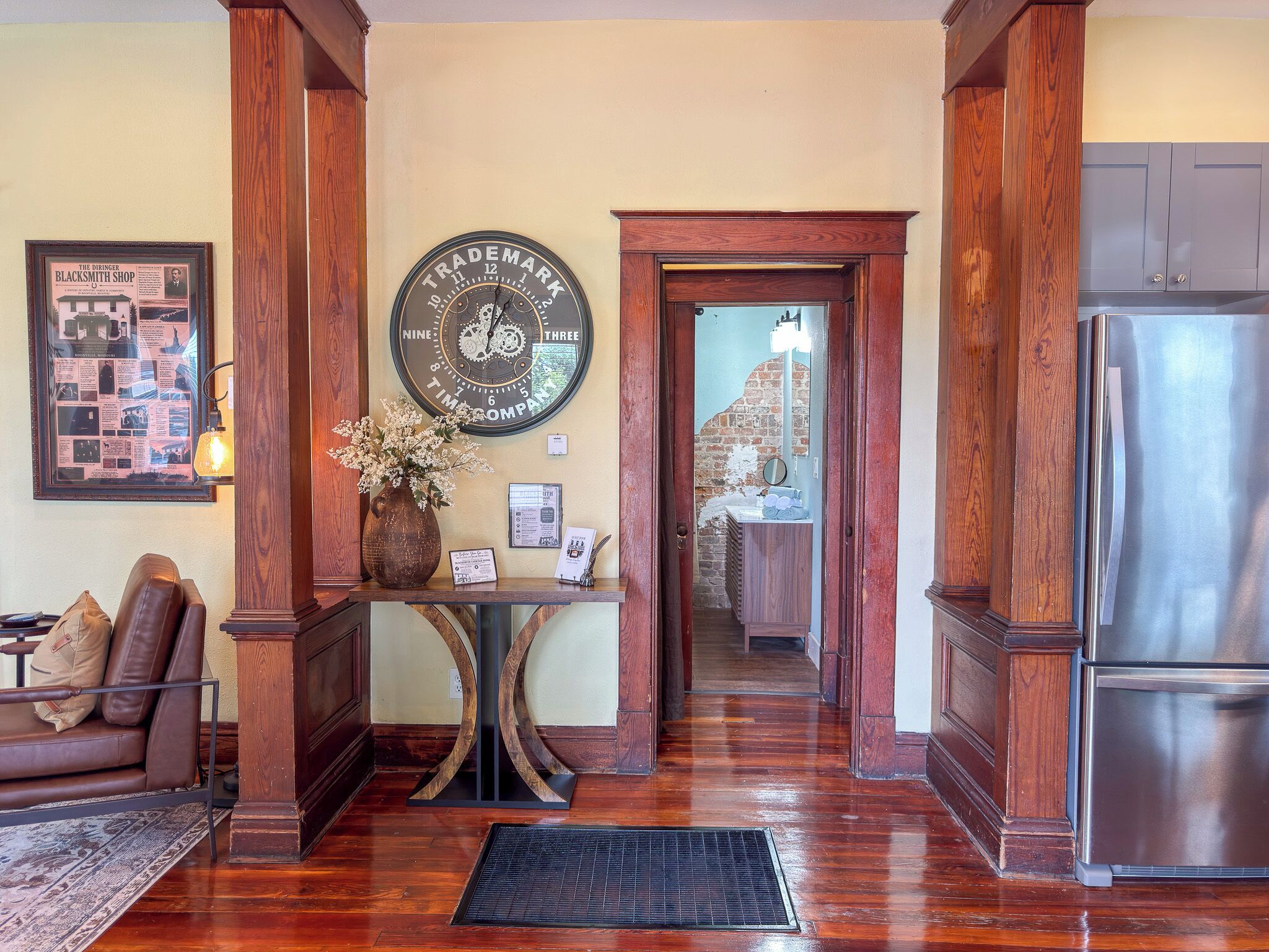 A wooden-floored hallway with a clock, a framed historical poster, a vase with flowers, and a refrigerator.