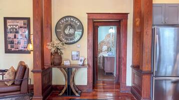 A wooden-floored hallway with a clock, a framed historical poster, a vase with flowers, and a refrigerator.