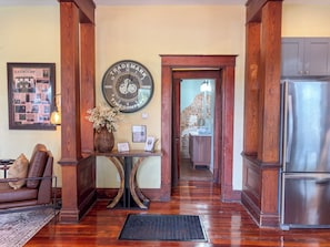 A wooden-floored hallway with a clock, a framed historical poster, a vase with flowers, and a refrigerator.