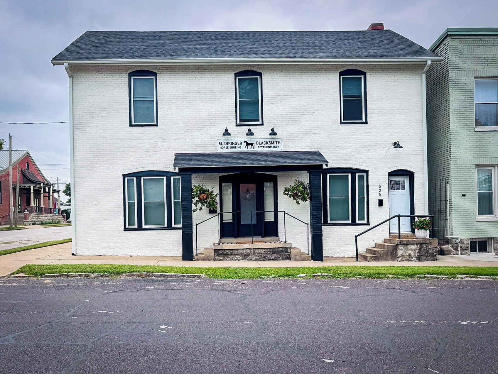 A two-story white building with a black front door and windows, a small porch with steps, and a sign above the door.