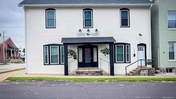 A two-story white building with a black front door and windows, a small porch with steps, and a sign above the door.