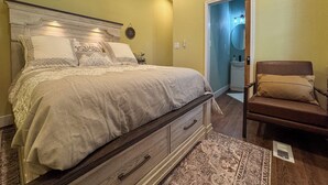 Een slaapkamer met een bed, een stoel, een nachtkastje, een dressoir en een deur naar een badkamer.