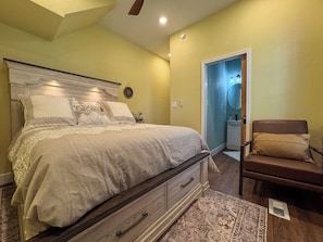 Ein Schlafzimmer mit Bett, Sessel, Nachttisch, Kommode und einer Tür zu einem Badezimmer.