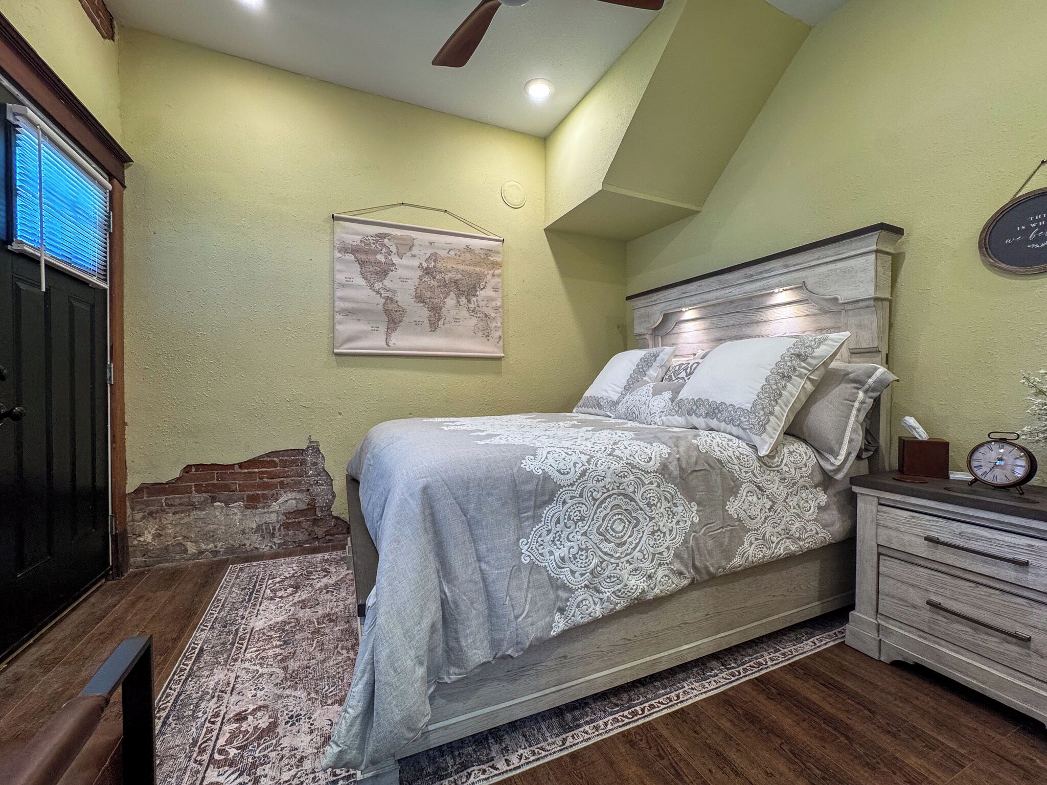 A bedroom with a bed, a nightstand, a clock, a world map on the wall, and a window with blinds.