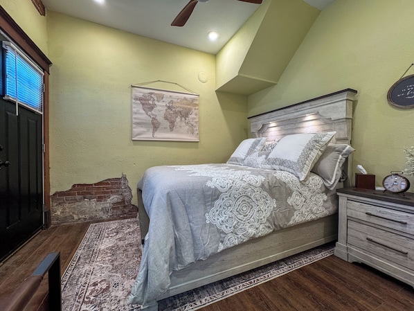 A bedroom with a bed, a nightstand, a clock, a world map on the wall, and a window with blinds.