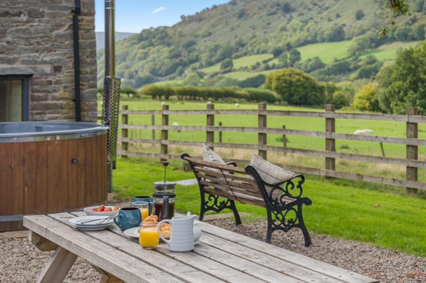 A wooden picnic table with breakfast items, a bench, and a hot tub in a scenic outdoor setting.