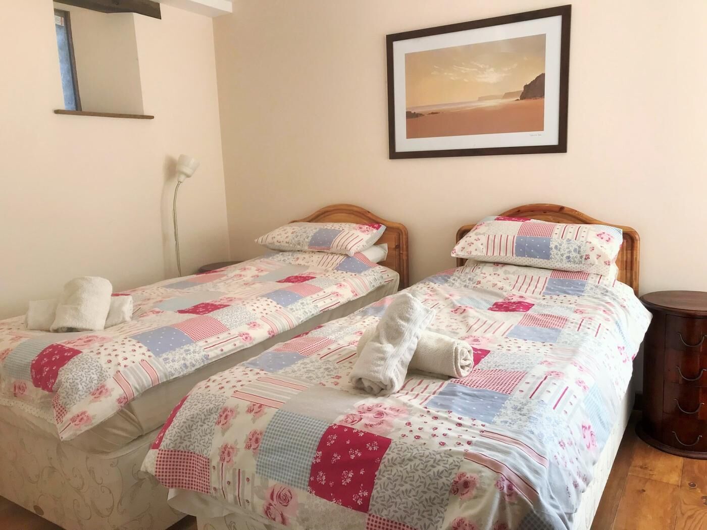 A room with two single beds, each with a patterned bedspread and pillows, a wooden headboard, a framed picture on the wall, and a bedside table with a lamp.