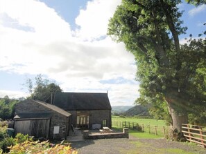 A rustic stone building with a sloped roof, a wooden fence, and a large tree in the foreground.