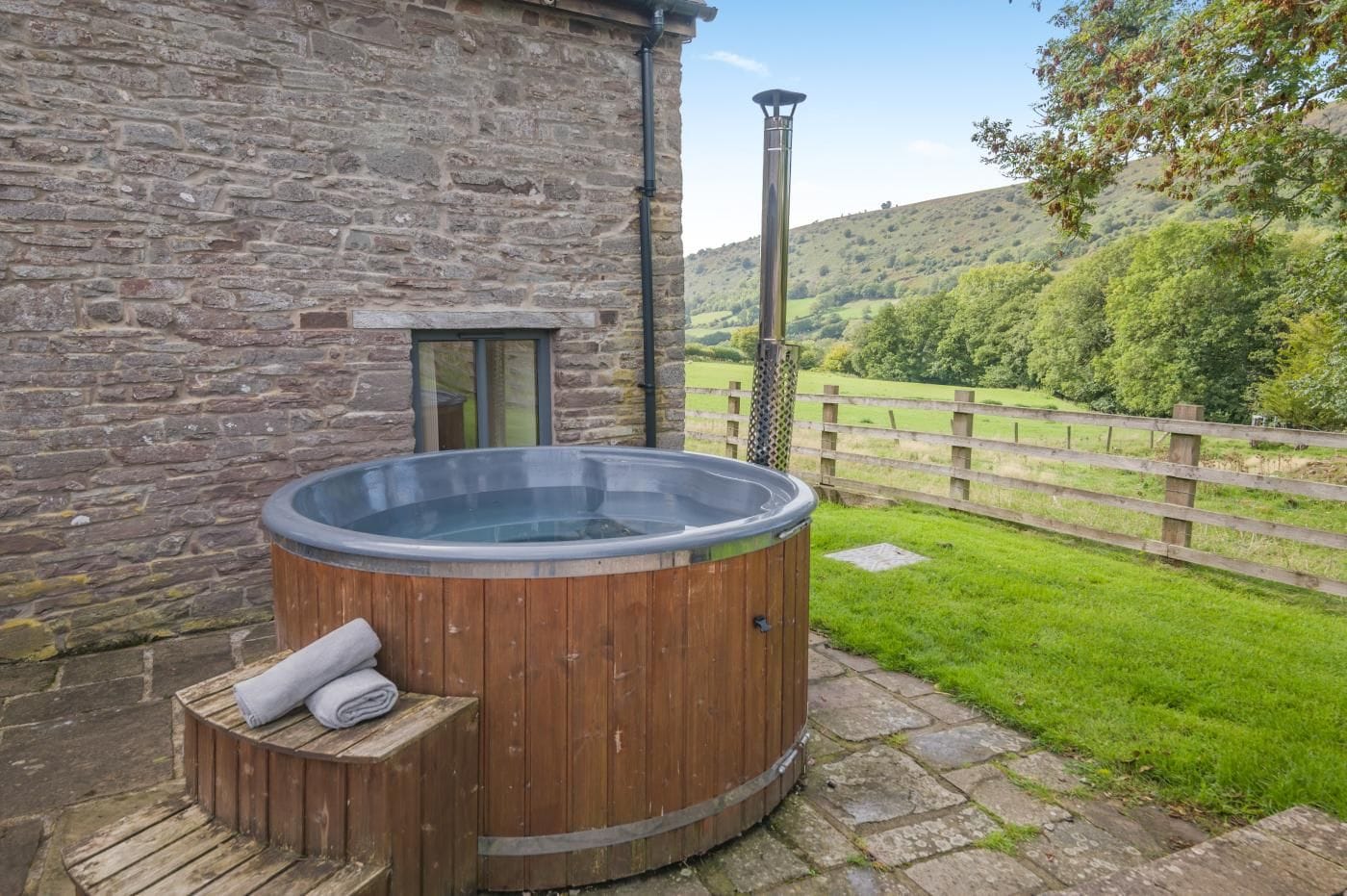 A wooden hot tub next to a stone building with a wooden deck and a wooden fence in the background.