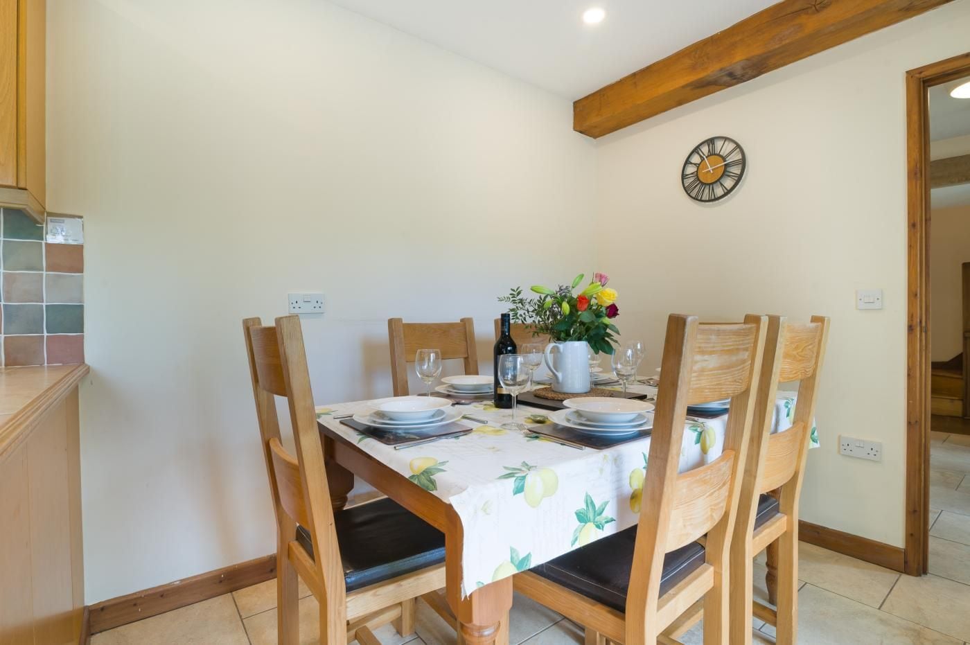 A dining area with a table set for four, wooden chairs, a clock on the wall, and a tiled backsplash.