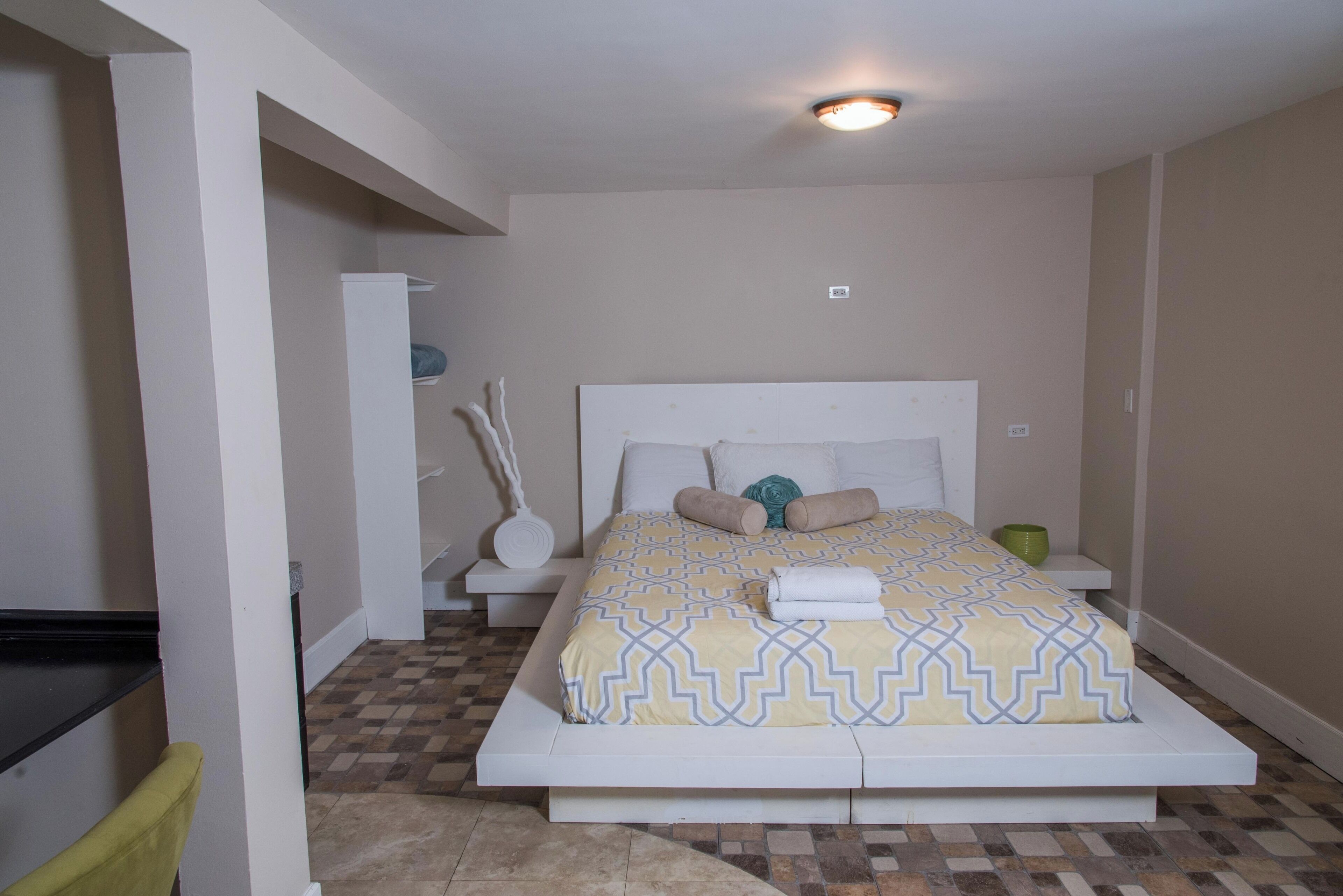 Un dormitorio con cama, mesita de noche, lámpara y toma de corriente.