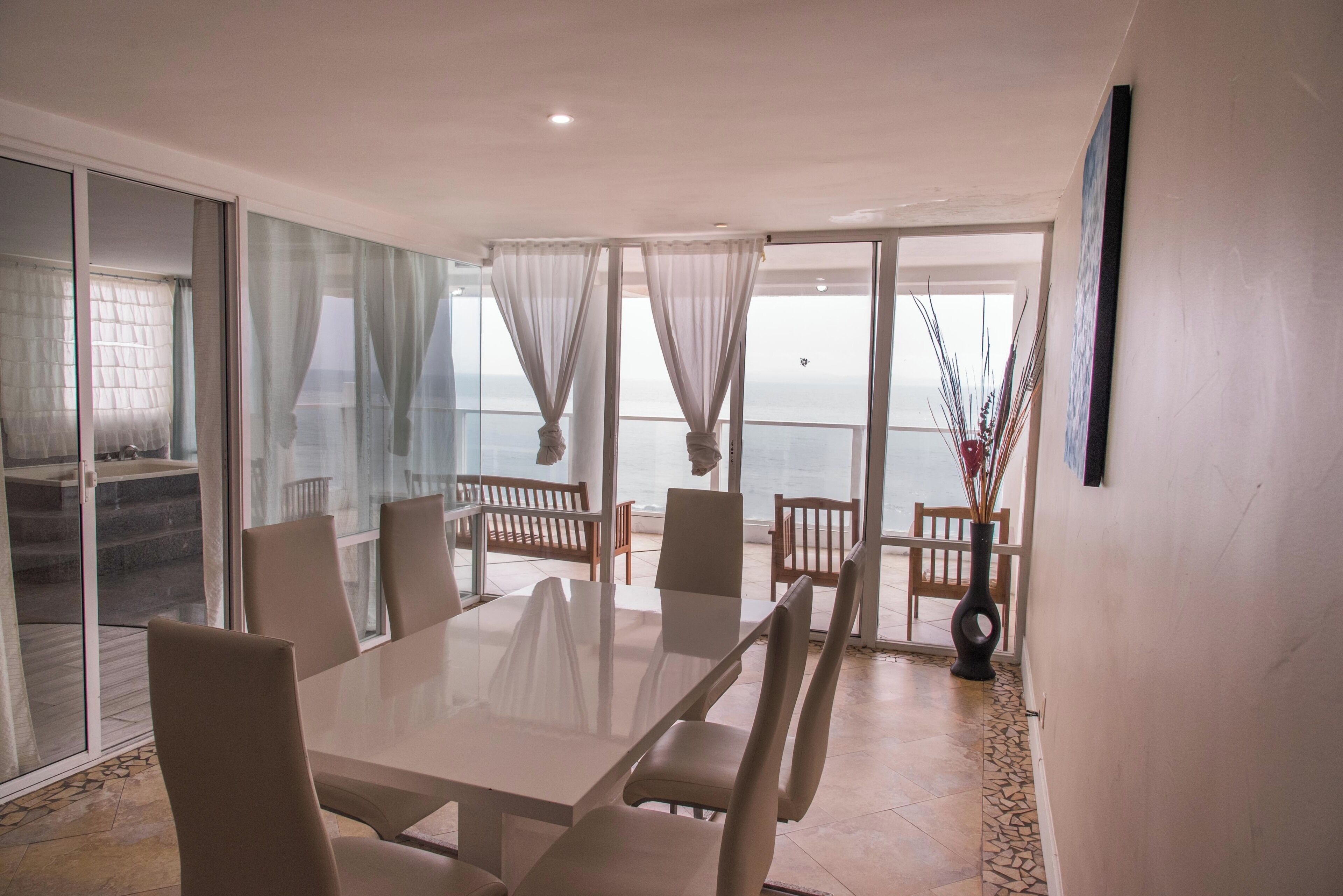 A spacious dining area with a large table and chairs, a view of the sea through large windows, and a balcony with a wooden bench.