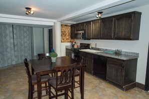 Una cocina con alacenas de madera oscura, encimera de granito, microondas integrado y lavavajillas. Hay una mesa de comedor con sillas y una planta en maceta.