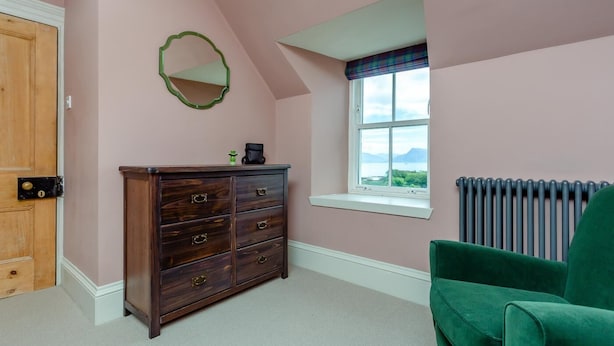 A room with a wooden dresser, a green chair, a window with a view, and a radiator. - 4 Bed in Ardvasar (oc-r43279) (Ardvasar)