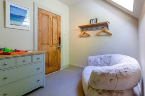 A room with a wooden door, a dresser with drawers, a cushioned chair, a shelf with hangers, and a framed picture on the wall.