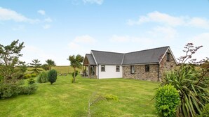 A two-story house with a stone facade and a white garage door, surrounded by a well-maintained lawn and various shrubs. - 3 Bed in Bryngwran (oc-y44816) (Bodedern)